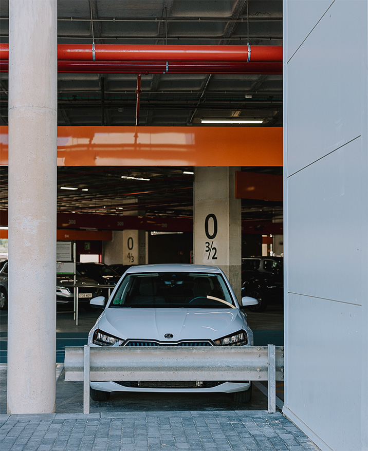 White car parked in a multi-level garage with concrete columns and orange overhead pipes.